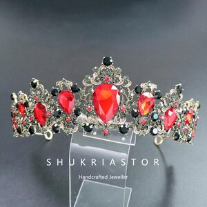 May include: An ornate crown with a vintage aesthetic. The crown features large, teardrop-shaped red gemstones and smaller black and red accents. The metalwork is a dark gold color, and the text "SHUKRIASTOR" is visible.
