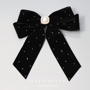 May include: A black velvet bow with scattered rhinestone accents. The bow features a central square-shaped pearl and rhinestone embellishment. The text "SHUKRIASTORE Handcrafted Jewellery" is visible at the bottom.