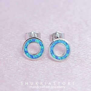 May include: Two silver stud earrings with a circular design. Each earring has a ring of blue and green iridescent stones set in a silver frame. The earrings are presented on a pink surface. The text "SHUKRIASTORE Handcrafted Jewellery" is at the bottom.