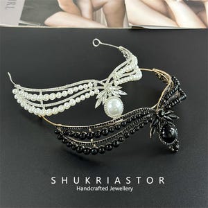 Può includere: Sono mostrate due fasce decorative, una bianca e una nera. La fascia bianca presenta perle e cristalli, mentre quella nera ha perline nere e una grande gemma nera. Il testo "SHUKRIASTOR Handcrafted Jewellery" è visibile.