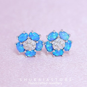 May include: A pair of flower-shaped stud earrings. Each earring features six oval, light blue opal stones surrounding a cluster of small, clear crystals. The earrings are set in a silver-colored metal. The text "SHUKRIASTORE Handcrafted Jewellery" is at the bottom.