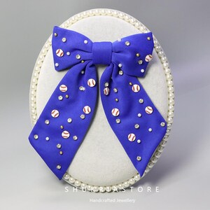 Peut inclure: Un nœud papillon bleu roi orné de motifs de baseball et d'ornements scintillants. Le nœud papillon présente de petits motifs de baseball et est orné de strass transparents et de perles dorées. Le nœud est présenté sur une base ovale blanche bordée de perles.