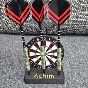 Könnte beinhalten: Drei Darts mit schwarz-rot gestreiften Flights werden vor einer Dartscheibe präsentiert. Die Darts haben silberne und grüne Akzente. Die Dartscheibe ist schwarz, rot, weiß und grün. Die Basis trägt den Namen "Achim" in Gelb.
