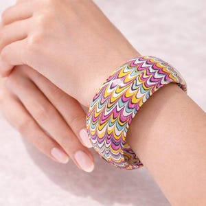 May include: A patterned bangle bracelet featuring a wavy design in pink, yellow, blue, and white. The bracelet is worn on a wrist, highlighting its bright colours. This accessory adds a vibrant touch to any outfit.