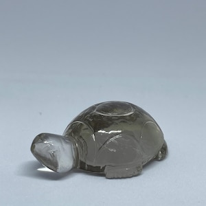 Puede incluir: Una figura de tortuga de piedra preciosa tallada y translúcida. La tortuga tiene un caparazón redondeado y una cabeza transparente. La piedra preciosa es de color gris ahumado. Es un objeto decorativo.