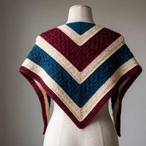 Op de afbeelding: Een handgemaakte sjaal in V-vorm, getoond op een mannequin. De sjaal heeft een textuursteekpatroon en een kleurblokontwerp met bordeauxrode, crèmekleurige en blauwgroene strepen. De randen zijn afgewerkt met een bordeauxrode rand.