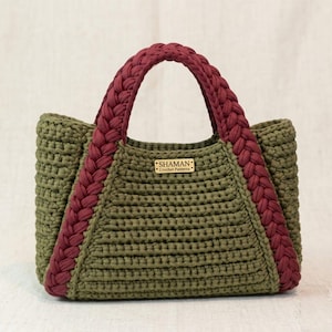 Puede incluir: Bolso tote de ganchillo hecho a mano en verde oliva con detalles y asas trenzadas en color burdeos. El bolso tiene forma rectangular y una pequeña etiqueta de madera con la inscripción "SHAMAN Crochet Patterns".