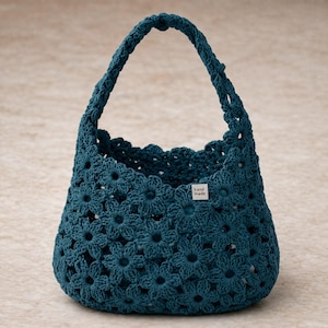 Könnte beinhalten: Türkisfarbene Häkeltasche mit floralem Muster. Die Tasche hat eine runde Form mit Henkel und einem gewellten oberen Rand. Ein kleines Etikett mit der Aufschrift "hand made" ist an der Tasche befestigt.