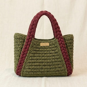 Puede incluir: Bolso tote de ganchillo hecho a mano en verde oliva con asas y ribetes trenzados en burdeos. El bolso tiene forma rectangular y una pequeña etiqueta de madera que dice "SHAMAN Crochet Patterns".