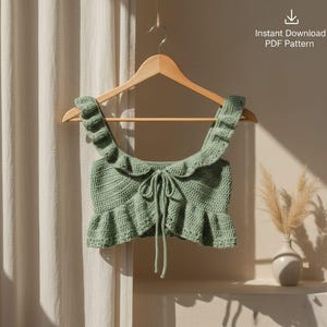 May include: A sage green crochet top with ruffled edges and a tie closure. The top is displayed on a wooden hanger against a neutral backdrop. The text "Instant Download PDF Pattern" is visible in the upper right corner.