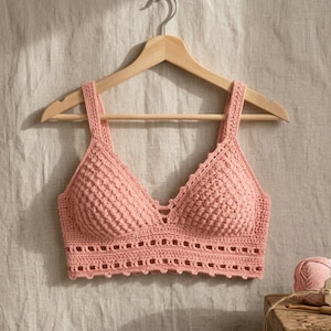 Puede incluir: Un top de ganchillo hecho a mano de color melocotón con un diseño de cuello en V, colgado de una percha de madera. El top presenta un patrón de puntadas texturizadas y bordes decorativos. Se ve un ovillo de hilo a juego y una etiqueta.