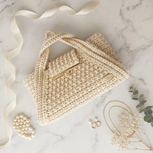 May include: Cream-coloured crochet handbag with a gold-tone clasp. The bag features a textured stitch pattern and a small wooden tag. Accessories include a pearl bracelet, gold earrings, and a necklace, on a marble surface.
