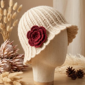 May include: A cream-coloured crocheted bucket hat featuring a ribbed texture and a prominent, layered dark red flower appliqué attached to the side. The hat is displayed on a neutral-toned mannequin head, showcasing its soft, knit construction.