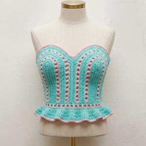 May include: A turquoise crochet top with a sweetheart neckline and a ruffled hem. The top features pink accents along the edges and vertical stripes. The garment is displayed on a mannequin.