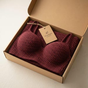 May include: A burgundy crochet bra and matching fabric are presented inside an open cardboard box. A small tag with the text "SHAMAN CRAFT" hangs from the bra. The bra features a classic bralette design with adjustable straps.