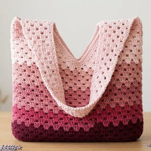 Può includere: Borsa tote all'uncinetto fatta a mano in una sfumatura di rosa e bordeaux. La borsa presenta un motivo a punto nonna e ampie cinghie. La borsa è su una superficie di legno.