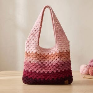 May include: A handmade crochet tote bag in shades of pink, peach, and burgundy. The bag features a textured stitch pattern and a single shoulder strap. Several balls of yarn and a crochet hook are on the table.