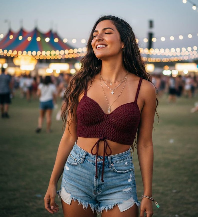 May include: A burgundy crochet crop top with spaghetti straps and a tie front. The top is paired with light wash denim shorts with frayed edges. The image is taken outdoors with a blurred background of a crowd and string lights.