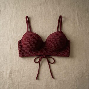 May include: A burgundy crochet top with adjustable straps and a tie closure at the front. The handmade top features a textured stitch pattern and a classic bra silhouette. The garment is displayed on a neutral background.