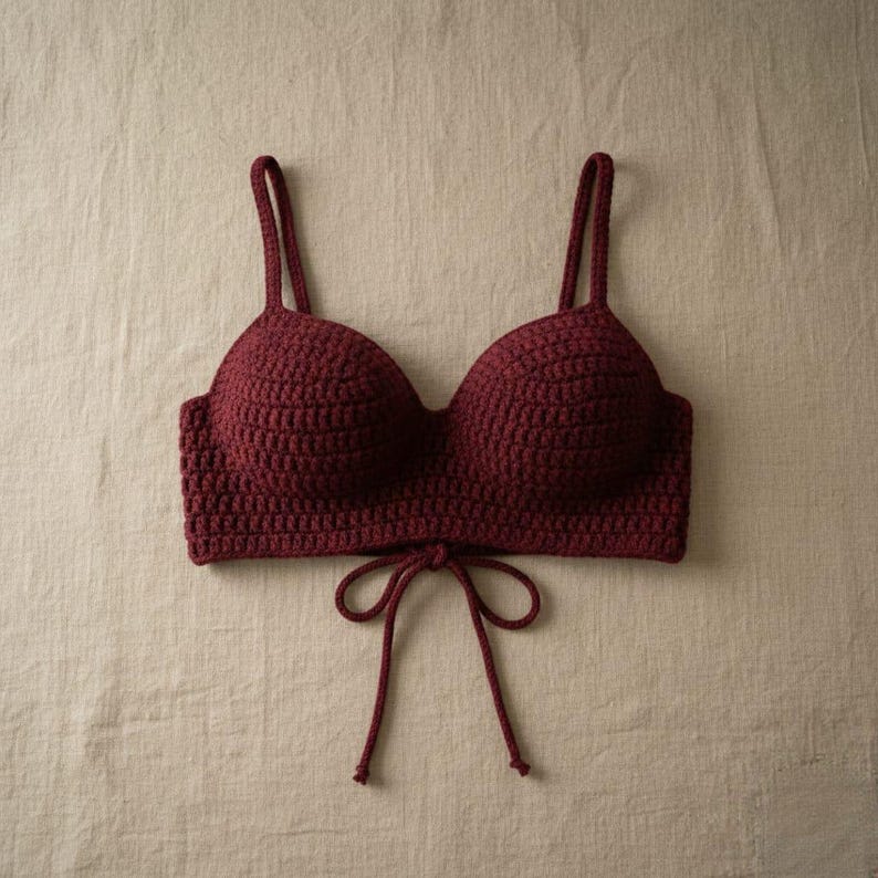 May include: A burgundy crochet top with adjustable straps and a tie closure at the front. The handmade top features a textured stitch pattern and a classic bra silhouette. The garment is displayed on a neutral background.