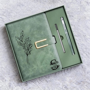 May include: Sage green journal and pen set in a gift box. The journal features a gold clasp and a black floral design. A silver pen and a refill are included. The journal has the name "Sopia" printed on it.