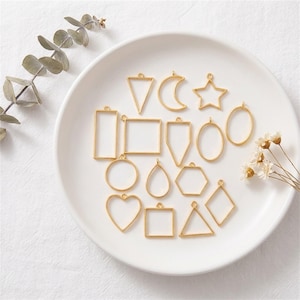 May include: A collection of gold-colored metal pendant frames in various shapes, including triangles, squares, hearts, and stars, arranged on a white plate. The shapes are open in the center, suitable for resin or other fillings.