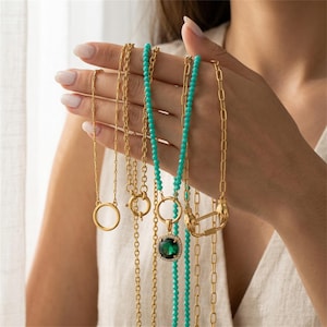 Puede incluir: Una colección de collares dorados con varios estilos de cadena, incluyendo una cadena fina con un colgante circular, una hebra de cuentas turquesas y una cadena dorada con un colgante cuadrado que sostiene una gran piedra verde esmeralda.