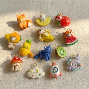 May include: A collection of twelve ceramic charms featuring various designs including a fox, a cat in a banana, a strawberry fox, a pineapple cat, a mushroom house, a blue cat, a kiwi slice, a watermelon red panda, a cloud, a rocket, and a snail.