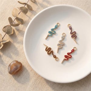 May include: Five snake-shaped charms with gold-colored metal and colorful rhinestones. The charms are in shades of blue, white, purple, brown, and red. The charms are on a white plate.