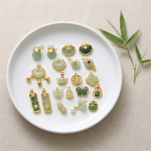 May include: A collection of jade pendants in various shapes, including bags, butterflies, and gourds, with gold-colored accents. The pendants are arranged on a white plate, with a sprig of bamboo in the background. The jade is a light green color.