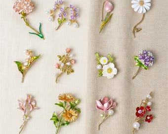 May include: A collection of twelve gold-tone metal brooches shaped like various flowers and plants, including daisies, tulips, hydrangeas, and lilies, featuring enamel details in pink, white, purple, red, and yellow with small faux pearl accents.