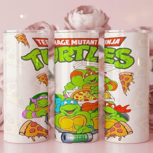 May include: Three white insulated tumblers featuring the Teenage Mutant Ninja Turtles. Each tumbler has a different design, including the TMNT logo, pizza slices, and the turtles themselves. The tumblers are cylindrical and have a silver lid.