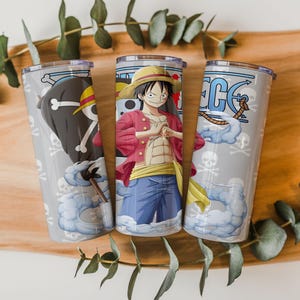 May include: Three stainless steel tumblers with One Piece anime character designs. Each tumbler features a different character illustration, including Luffy, and pirate-themed graphics. The tumblers have clear lids and are set against a wooden background.