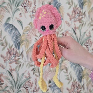 May include: A handmade crochet octopus toy featuring a textured pink head with yellow accents, large black safety eyes, and dangling tentacles in shades of coral pink and bright yellow. The soft yarn creature has a rounded top and long limbs.