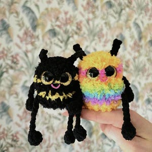 May include: Two handmade crochet creatures. One is black with yellow accents and a pink mouth, the other is yellow with rainbow stripes. Both have large, glittery eyes and black limbs.