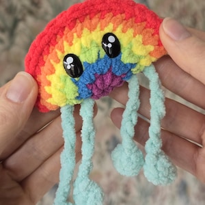 May include: A handmade, colourful crochet jellyfish toy. The jellyfish has a rainbow-coloured head with large, black, googly eyes and light blue tentacles with pom-pom ends. The toy is held in two hands.