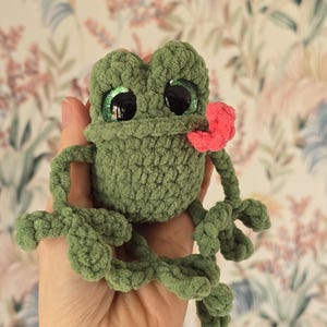 May include: A green crochet frog with large black eyes and a pink tongue. The frog is handmade, with detailed features and a plush texture. It is held in a hand, highlighting its whimsical design. A charming decorative item or toy.