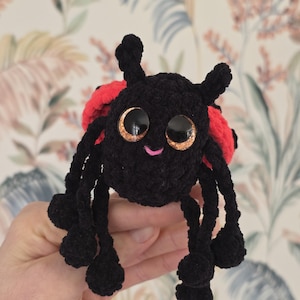 Puede incluir: Una mariquita negra hecha a mano con ganchillo, con alas rojas y ojos grandes y brillantes. La mariquita tiene seis patas negras y dos antenas. Es un juguete blando.