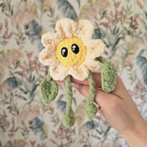Puede incluir: Una flor de ganchillo con pétalos de color crema y un centro amarillo brillante. La flor presenta dos grandes ojos de seguridad negros con reflejos blancos, dos hojas verdes de ganchillo y dos tallos verdes que terminan en pequeños capullos.