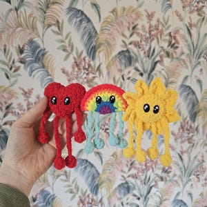May include: Three crocheted plush toys: a red heart, a rainbow, and a yellow sun. Each has black button eyes and dangling legs. The heart and sun have four legs, while the rainbow has four blue legs. The toys are handmade and colorful.