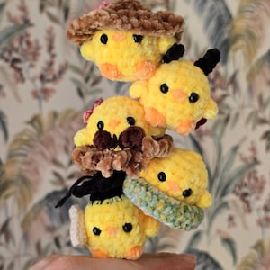 May include: A stack of five yellow plush chick toys with black eyes, orange beaks, and various accessories. The accessories include a brown hat, a pink flower, a black bow, and a green and white scarf. The toys are handmade and have a soft, fuzzy texture.