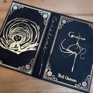 Puede incluir: Libro abierto con cubierta negra y detalles dorados. El lado izquierdo presenta una ilustración dorada de una persona en un túnel. El lado derecho tiene el título "Coraline" y el autor "Neil Gaiman" en dorado. Bordes decorativos.
