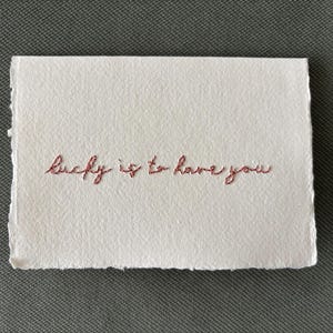 Può includere: Un biglietto rettangolare bianco con bordi sfrangiati presenta la frase ricamata "lucky is to have you" in filo marrone rossiccio. Il biglietto è posto su uno sfondo grigio strutturato.