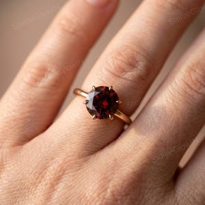 Könnte beinhalten: Ein Ring mit einem runden, tiefroten Edelstein in einer polierten Roségoldfassung. Der Stein wird von sechs Krappen gehalten und besitzt eine facettierte Oberfläche, die das Licht reflektiert. Die Ringschiene ist schmal, glatt und metallisch.