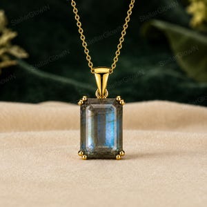May include: A gold necklace featuring a rectangular emerald-cut labradorite pendant with a blue iridescent sheen. The gemstone is held in a four-prong setting attached to a delicate gold chain, showcasing a polished metallic finish and clear stone facets.