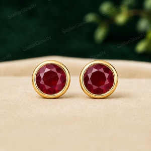May include: A pair of stud earrings featuring round, faceted deep red rubies set in polished yellow gold bezels. The stones exhibit a brilliant cut with multiple facets that reflect light, encased in a smooth, circular gold metal frame.