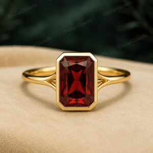 May include: A gold ring featuring a large, rectangular, emerald-cut deep red garnet gemstone set in a smooth bezel setting. The band has a split shank design where the metal arms meet the central stone setting, creating a polished gold finish.