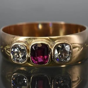 May include: A gold ring featuring three gemstones: two clear, square-cut stones flank a central, square-cut ruby. The ring's band is wide and polished, reflecting light. The ring is displayed on a reflective surface.