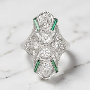 May include: A silver ring with a geometric design, featuring multiple round and square-cut diamonds. Emerald-colored stones are set on the sides. The ring has an intricate filigree pattern.