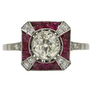 Peut inclure: Bague en platine ornée d'un diamant central clair et rond. Le diamant est entouré d'un cadre carré de petits rubis carrés et de diamants d'accent. La bague présente un design Art Déco classique.